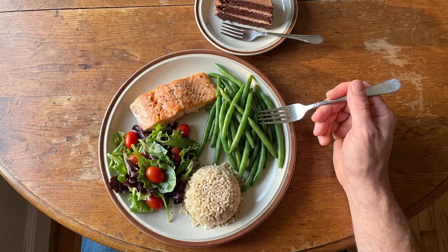A dinner plate with salmon, green beans, salad and brown rice, and a thin slice of chocolate cake waiting on a saucer