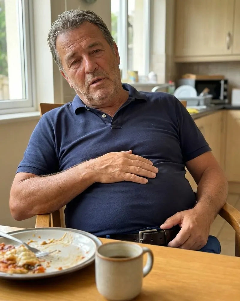 Ray at the breakfast table after eating, hand on his bloated stomach under a tight navy polo, looking worn out