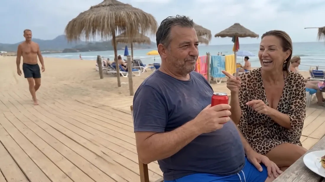 A 64-year-old man in a tight navy t-shirt holding a soda at a beach bar, grinning, while his wife laughs and points at a fit man walking by on the sand