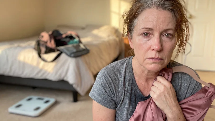 A woman in her fifties, still sweaty from the gym, clutches a pink bridesmaid dress with a bathroom scale on the floor behind her