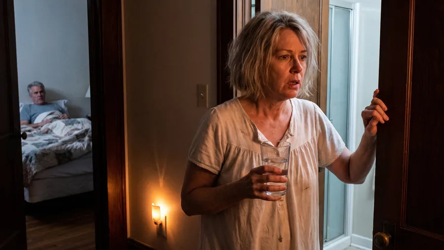 At 3 a.m. a woman in a nightgown grips the door frame in a dark hallway, a glass of water in her hand, while her husband sits up in bed behind her